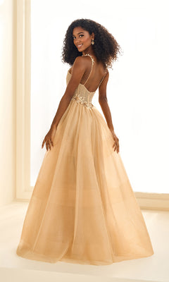 A woman in the Ellie Wilde EW36058 long prom dress—featuring shimmering light gold glitter tulle, thin straps, and a corset bodice—stands in a well-lit room, looking over her shoulder and smiling.