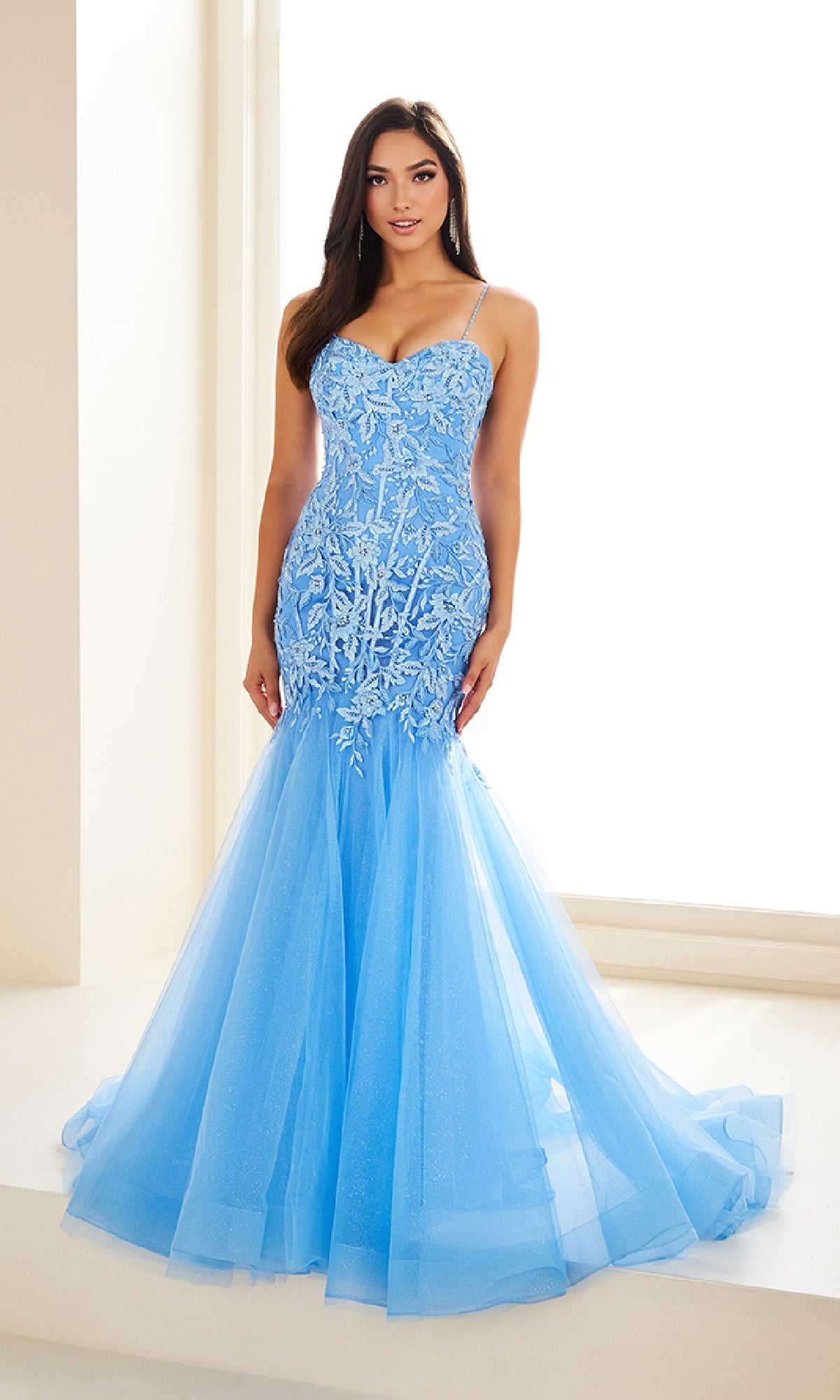 A woman stands indoors wearing the Ellie Wilde EW36062, a fitted light blue mermaid prom dress with a lace bodice, thin straps, and an adjustable lace-up back.