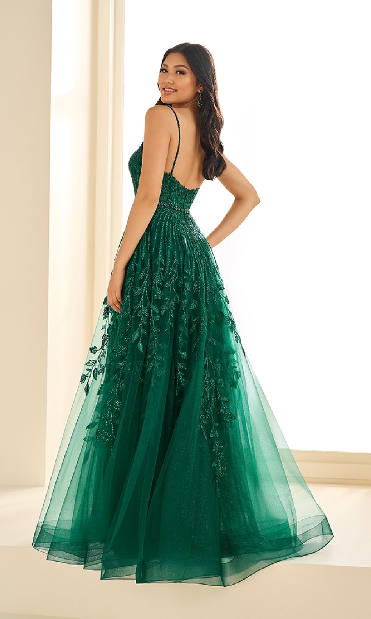 A woman in the Ellie Wilde EW36068 long prom dress—sleeveless, floor-length, with floral embroidery and glitter tulle—stands indoors, looking over her shoulder.