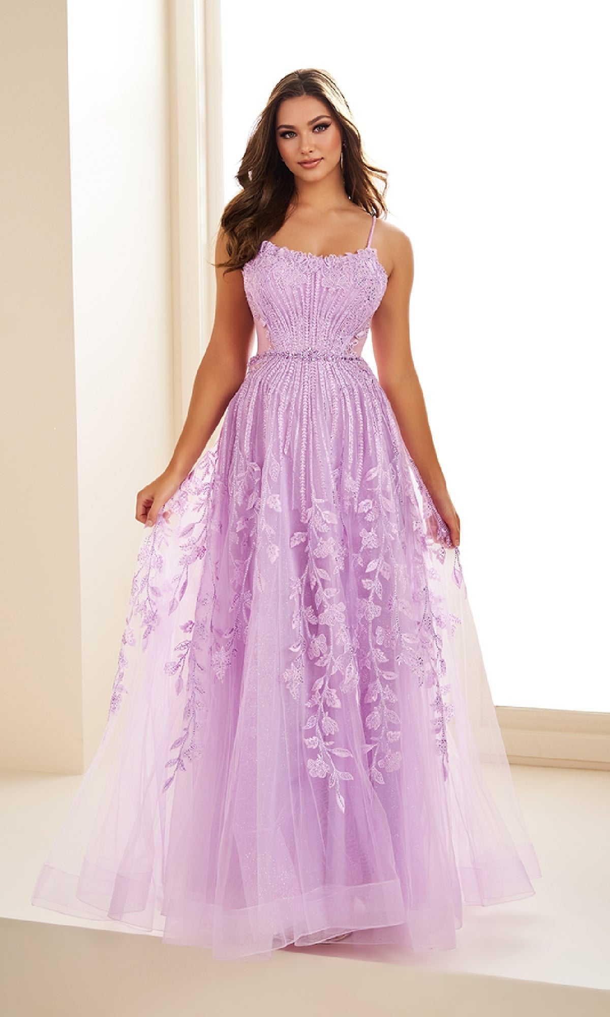 A woman stands indoors near a window, holding the sides of her skirt while wearing the Ellie Wilde EW36068 long prom dress featuring sleeveless design with lace and floral embroidery in lavender.