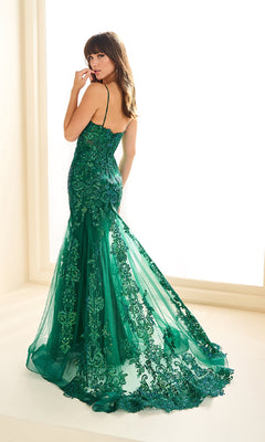 A woman in the Ellie Wilde EW36069, a fitted, floor-length green lace applique prom dress with thin straps, stands facing away from the camera in front of a window.