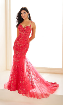 A woman stands indoors wearing the Ellie Wilde EW36069, a fitted, floor-length red glitter tulle mermaid prom dress with gold lace appliqué, thin straps, sheer corset bodice, and flared semi-sheer train.