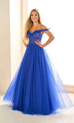 A woman poses indoors in the Ellie Wilde EW36071 long prom dress, an off-the-shoulder gown with a fitted bodice and glitter tulle skirt detailed with small beaded embellishments.