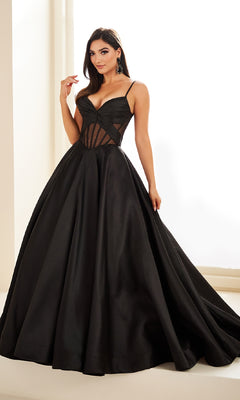 A woman poses indoors in the Sheer-Corset Prom Ball Gown: Ellie Wilde EW36073, a long black dress with pockets, fitted bodice, sheer panels, and sweetheart neckline. She gracefully holds the skirt with one hand.