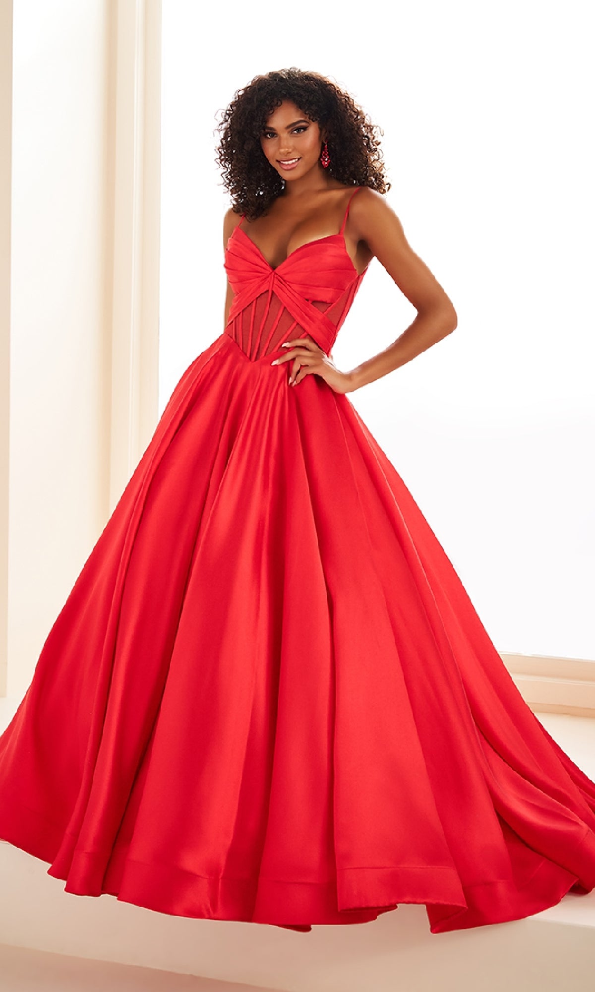 Woman in the Ellie Wilde EW36073 Sheer-Corset Prom Ball Gown, a vibrant red dress with a sweetheart neckline and fitted bodice, stands indoors smiling at the camera.