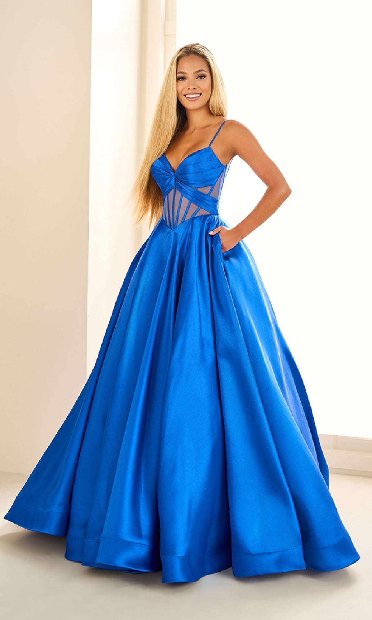 A woman in the Ellie Wilde EW36073 Sheer-Corset Prom Ball Gown, featuring vibrant blue color, pockets, a fitted bodice, sheer cutouts, and spaghetti straps, stands indoors by a window.