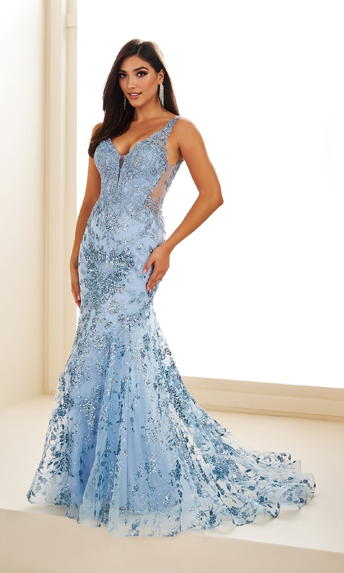 Indoors, a woman wears the Ellie Wilde EW36074 long prom dress—a fitted light blue mermaid gown with floral sequins, lace embroidery, and sheer side panels.