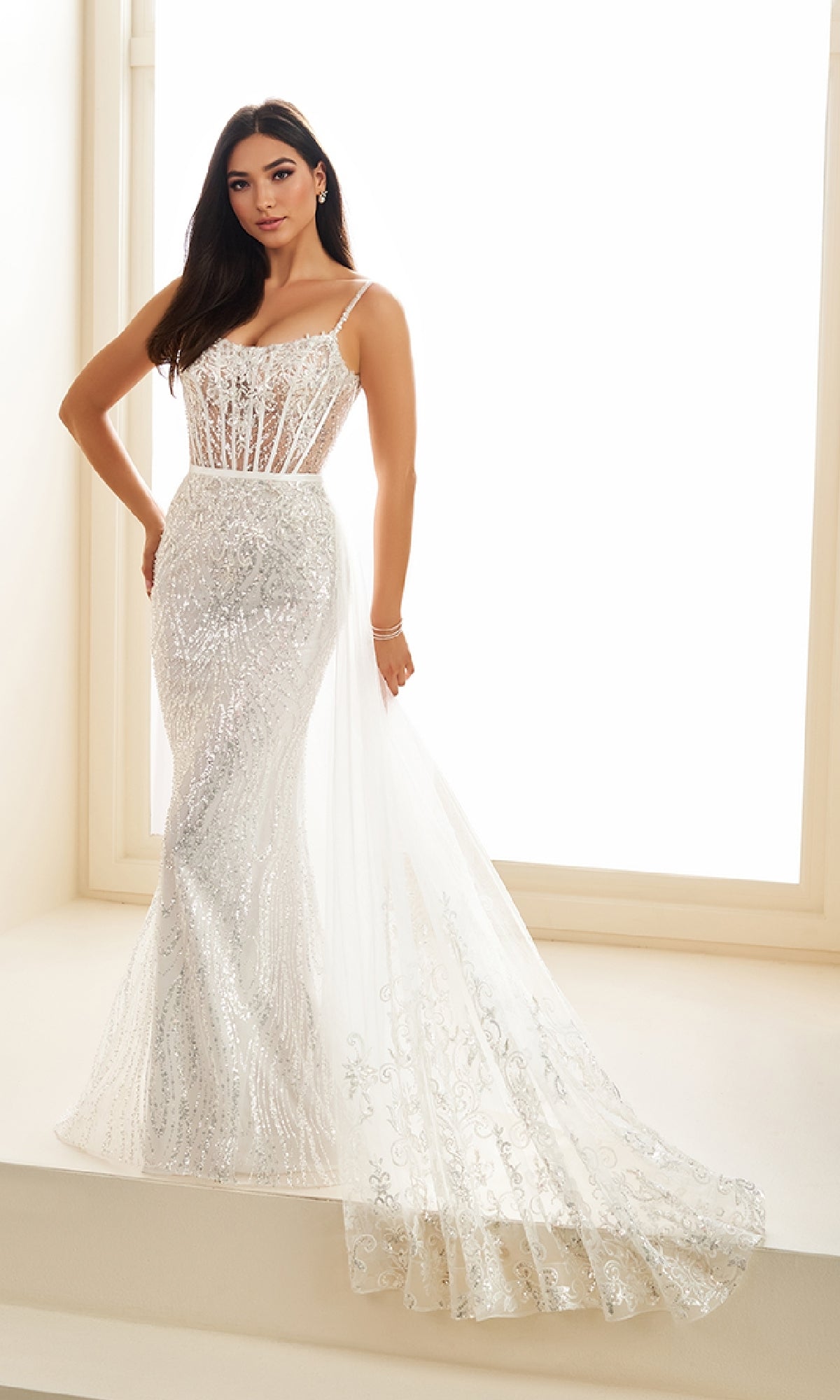 Woman indoors by a large window, wearing the Ellie Wilde EW36075— a long white fitted prom dress with embroidered tulle, sheer corset bodice, and thin straps.