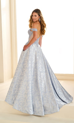A woman wearing the Ellie Wilde EW36078 long prom dress with a light blue embellished corset bodice and a patterned skirt stands facing backward, looking over her shoulder and smiling.