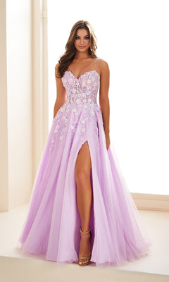 A woman stands indoors by a bright window wearing the Long Prom Dress: Ellie Wilde EW36079—a light purple gown with floral appliqué, thin straps, corset bodice, and a high slit.