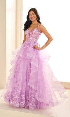 A woman stands indoors wearing the Ellie Wilde EW36081, a long lilac prom dress featuring floral lace appliqué, a fitted bodice with spaghetti straps, and a layered glitter tulle skirt.