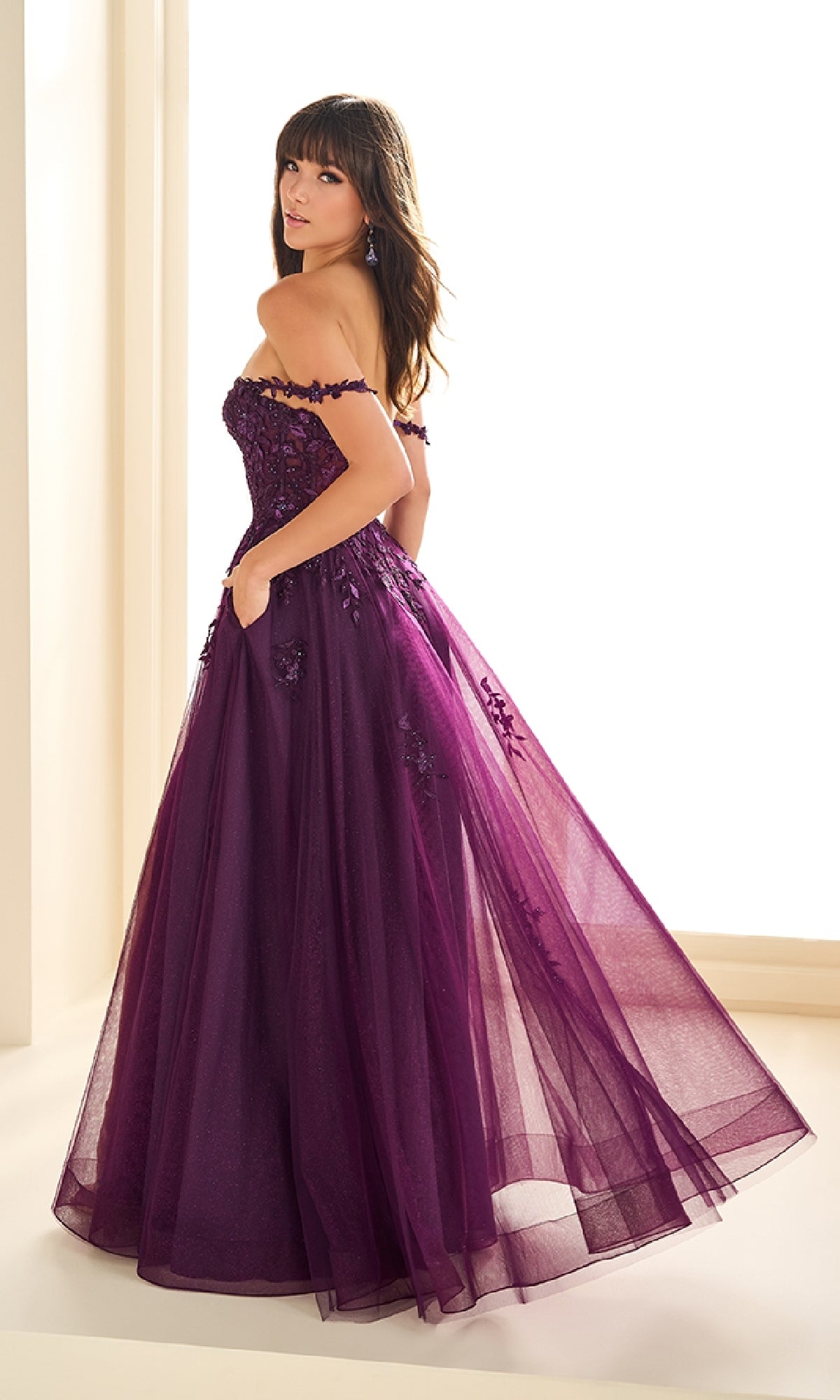 A woman in the Ellie Wilde EW36083 long prom dress—floor-length, off-the-shoulder, purple with floral accents and glitter tulle—stands in a brightly lit room, glancing over her shoulder.