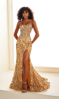 A woman stands near a large window indoors, wearing the Ellie Wilde EW36084 long prom dress—a gold sequin gown with a corset bodice, spaghetti straps, floor-length skirt, and thigh-high slit.