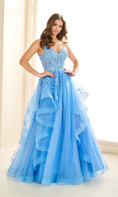 A woman stands in a room wearing the Ellie Wilde EW36085 long prom dress, featuring a light blue lace embroidered corset with floral details and a layered tulle skirt.