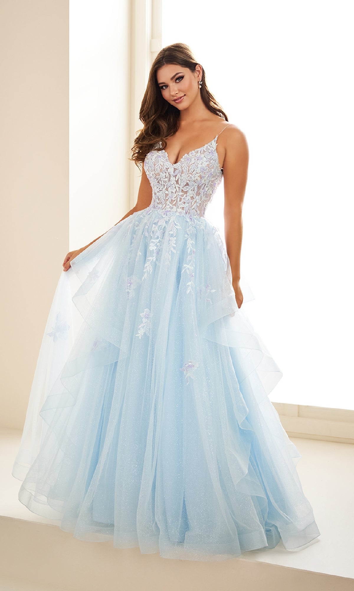 A woman stands by a window wearing the Ellie Wilde EW36085, a light blue glitter tulle prom gown featuring a fitted lace embroidered corset bodice with thin straps and a layered skirt.