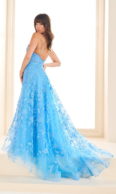 Woman wearing the Ellie Wilde EW36088 long prom dress in light blue, featuring a floor-length design, low back, thin straps, and elegant lace details, stands indoors looking over her shoulder.