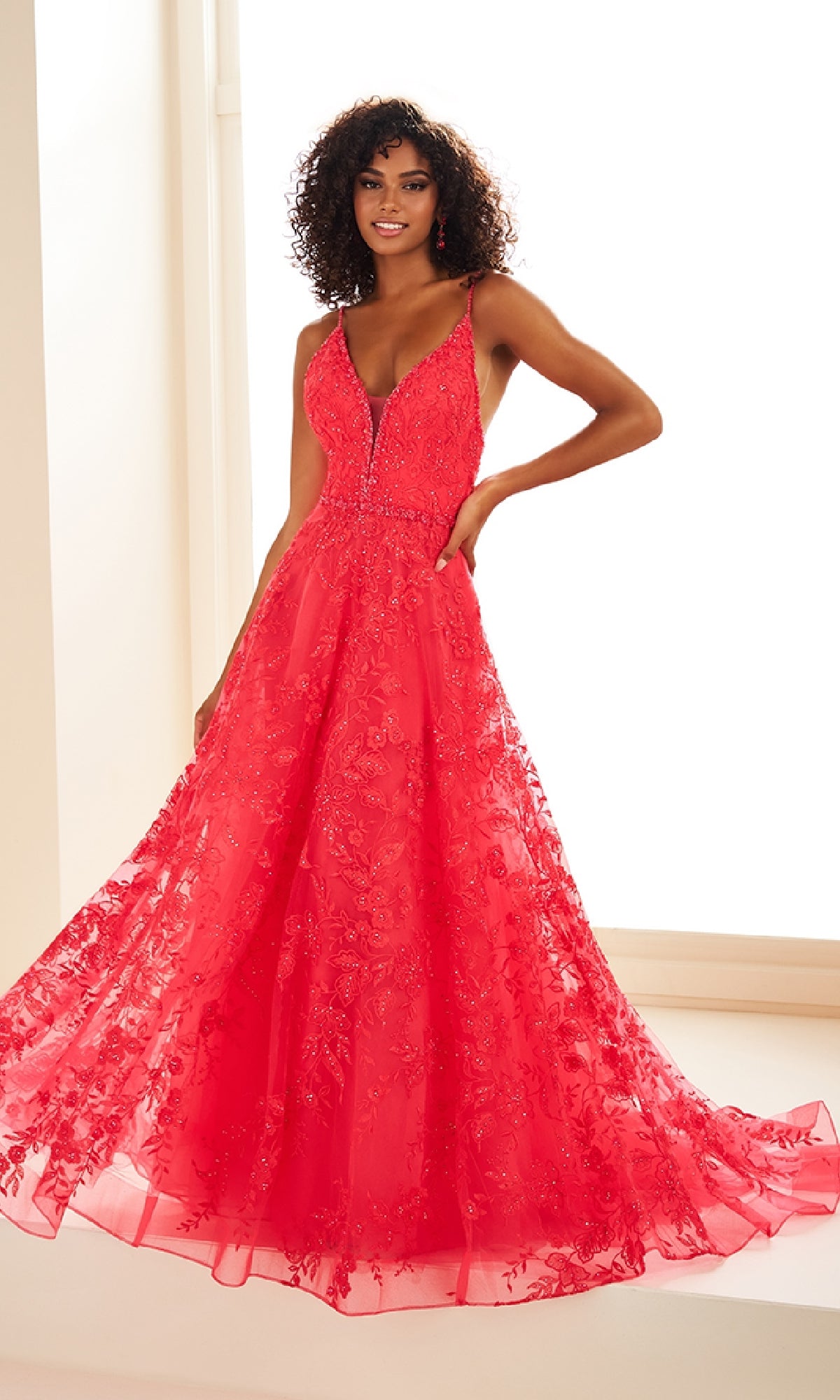 A woman stands in a brightly lit room wearing the Ellie Wilde EW36088 long prom dress—a bright red, sleeveless, floor-length gown with floral embroidery, an elegant a-line skirt, and pockets.