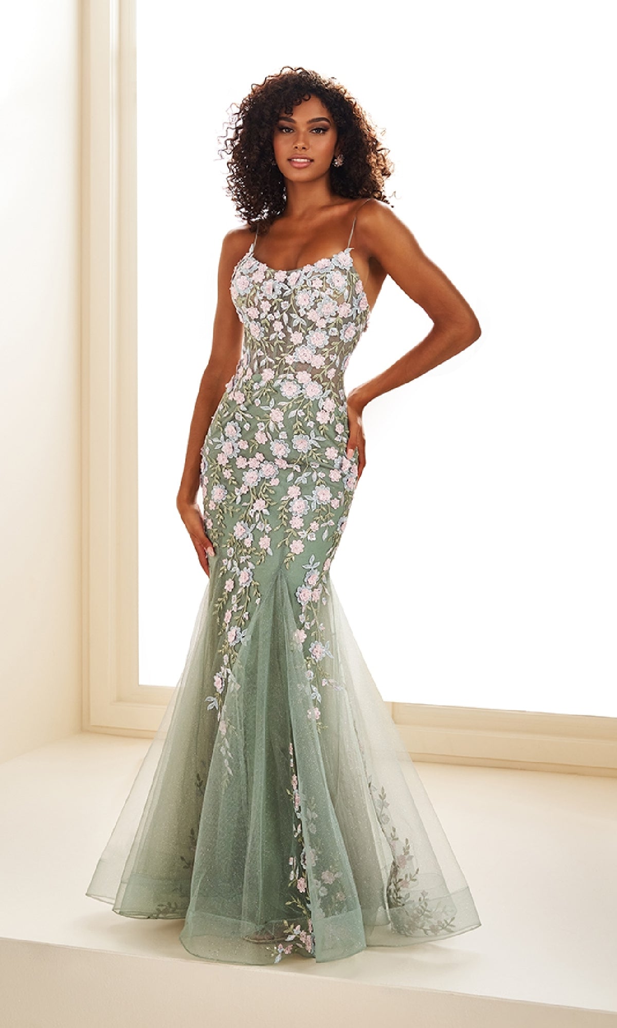 A woman stands by a bright window wearing the Ellie Wilde EW36090, a fitted sheer green long prom dress with floral lace appliques and thin straps.