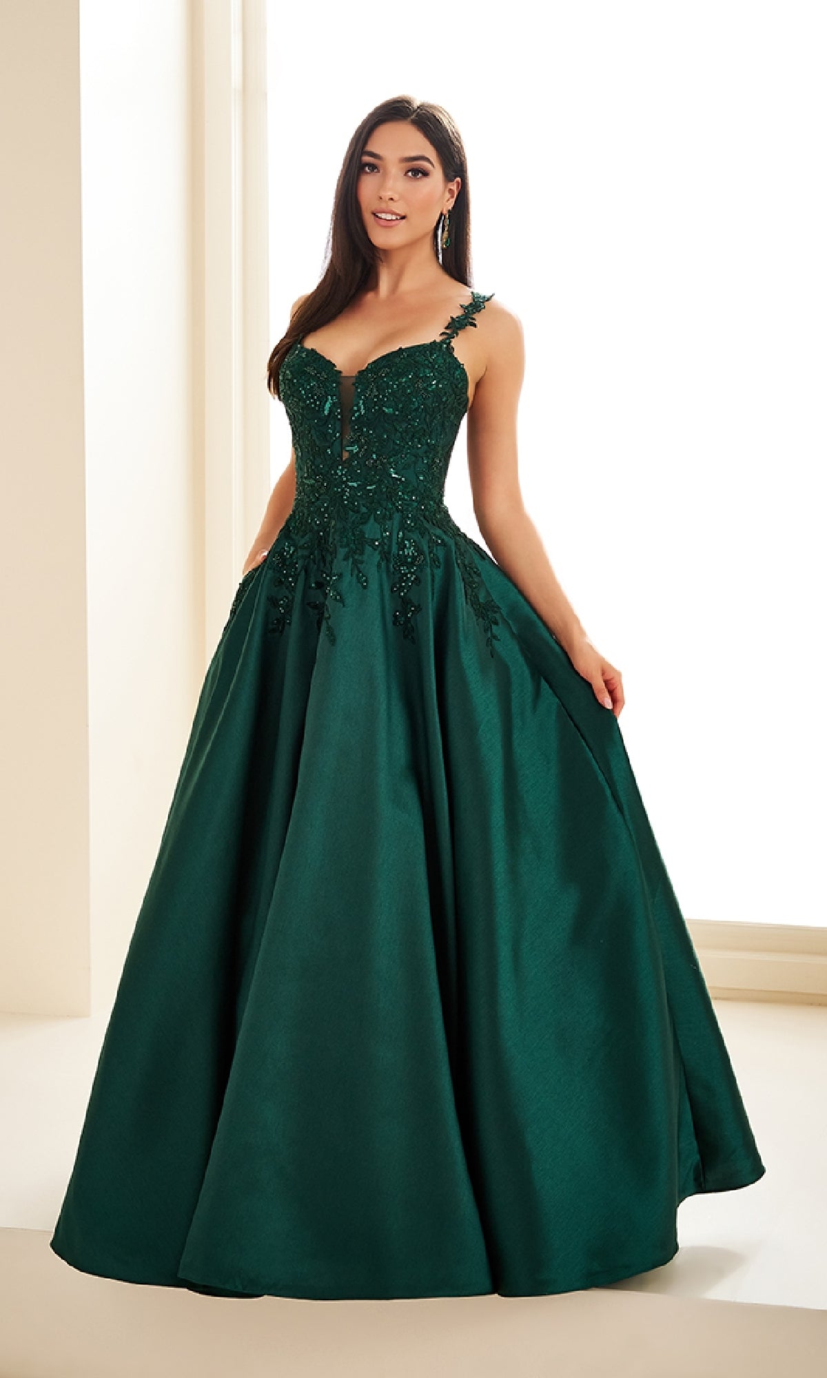 A woman stands indoors wearing the Ellie Wilde EW36094 long prom dress, a dark green floor-length gown with a fitted, embellished corset bodice and full skirt.