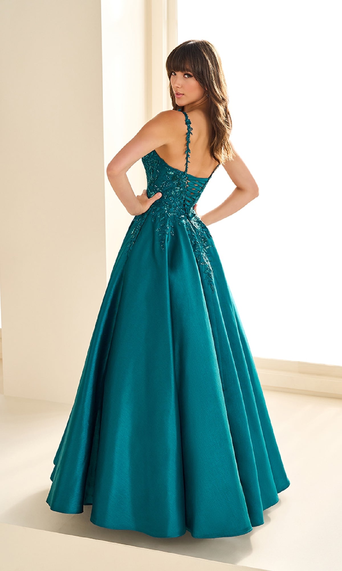 Woman wearing the Ellie Wilde EW36094 long prom dress in teal, featuring a corset bodice and lace detailing on brushed satin, standing indoors and looking over her shoulder.