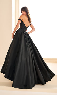 A woman stands near a bright window, looking over her shoulder while wearing the Ellie Wilde EW36100—a floor-length, off-the-shoulder prom ball gown with a fitted corset bodice.