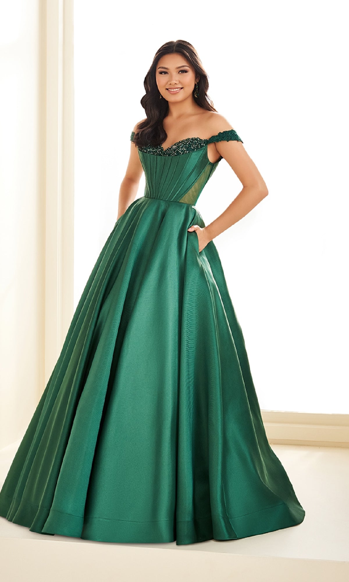 A woman in the Ellie Wilde EW36100 long prom dress, featuring an off-the-shoulder emerald green ball gown with a delicate lace corset bodice, stands indoors against a light background.