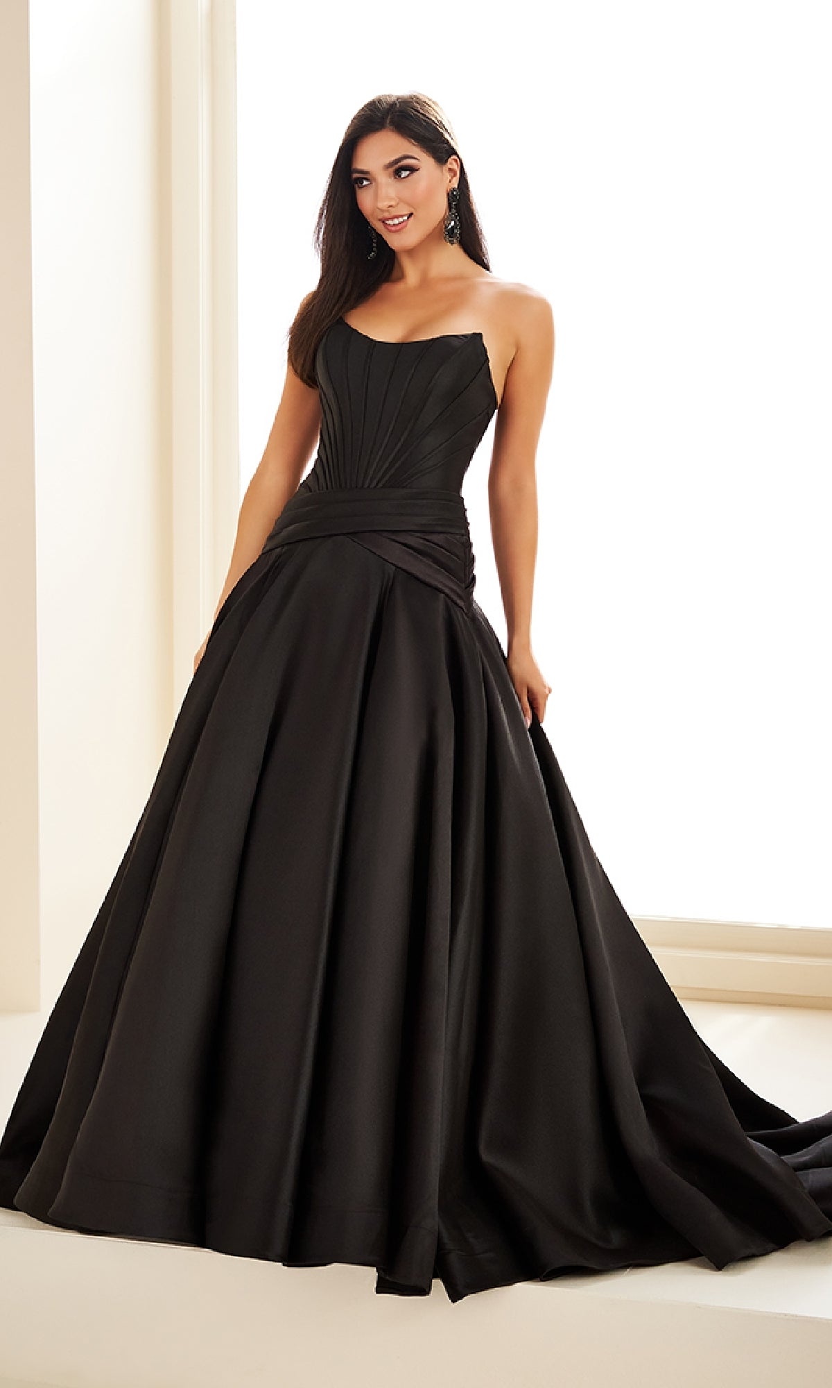 A woman stands indoors wearing the Ellie Wilde EW36105, a long prom dress with a strapless mikado ball gown silhouette, fitted corset bodice, and full floor-length skirt.