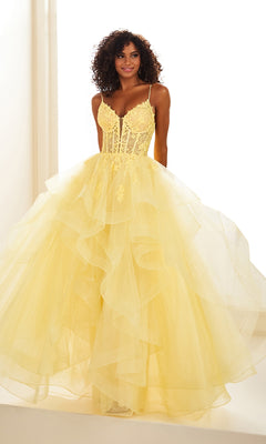 A woman with curly hair models the Ellie Wilde EW36203 Glitter Tulle Prom Ball Gown, featuring a yellow ruffled skirt, embroidered corset bodice, and thin straps, standing against a white background.