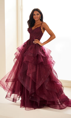 A woman stands indoors before a bright window, wearing the Ellie Wilde EW36203 Glitter Tulle Prom Ball Gown—sleeveless, dark red, with layered tulle skirt, an embroidered corset bodice, and adjustable lace-up back.