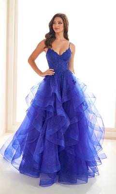 A woman stands in a bright room wearing the Ellie Wilde EW36203 Glitter Tulle Prom Ball Gown, featuring deep blue glitter tulle, thin straps, and a fitted embroidered corset bodice.