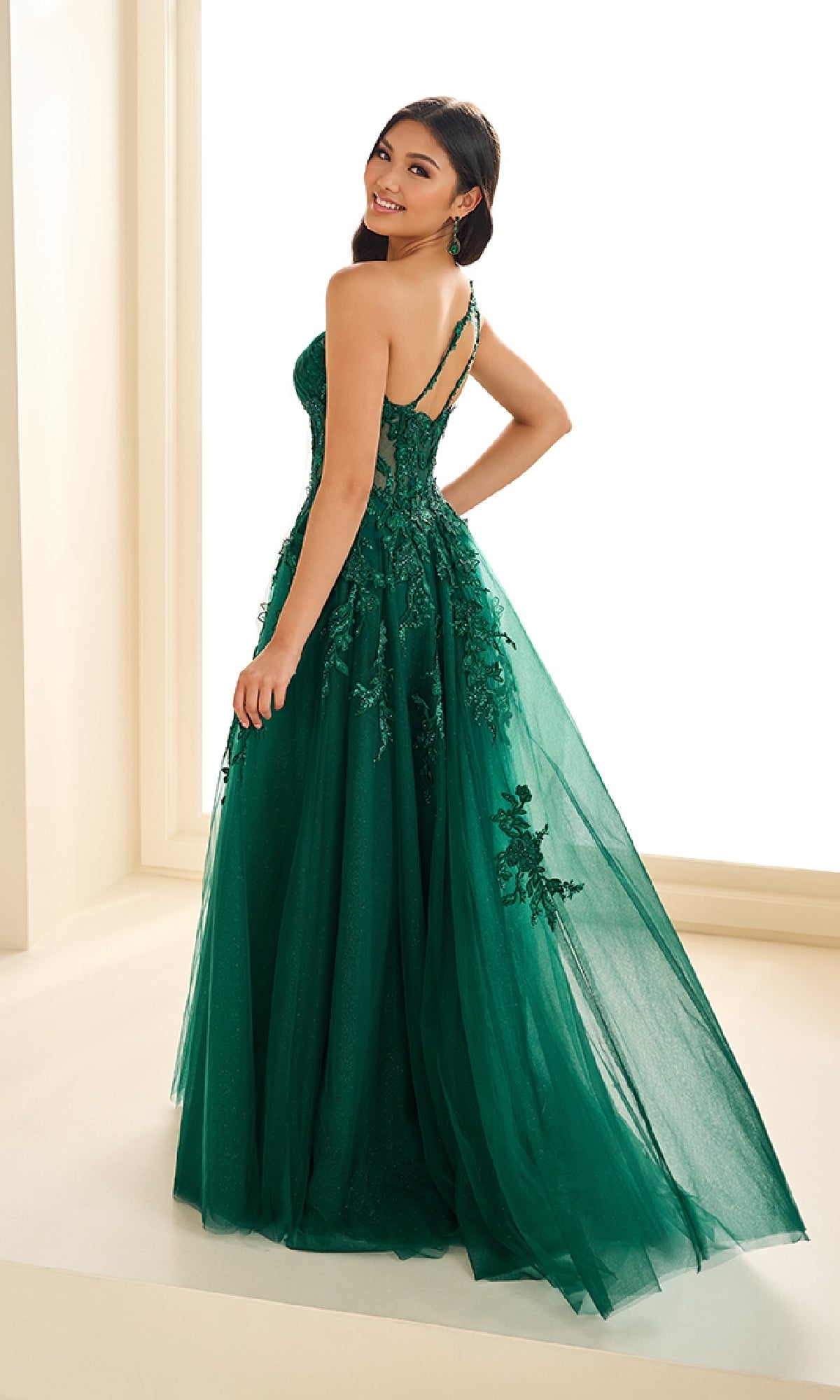 A woman stands indoors in the Ellie Wilde EW36207 long prom dress, featuring a floor-length green gown with floral embellishments and a corset bodice, looking over her shoulder and smiling.