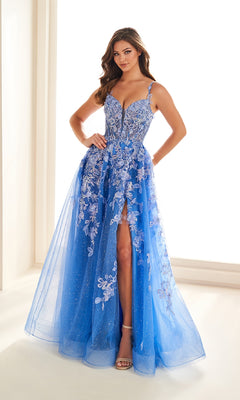 A woman wears the Ellie Wilde EW36208 long prom dress—blue glitter tulle with 3D flowers, corset bodice, spaghetti straps, and a thigh-high slit—standing indoors against a bright background.