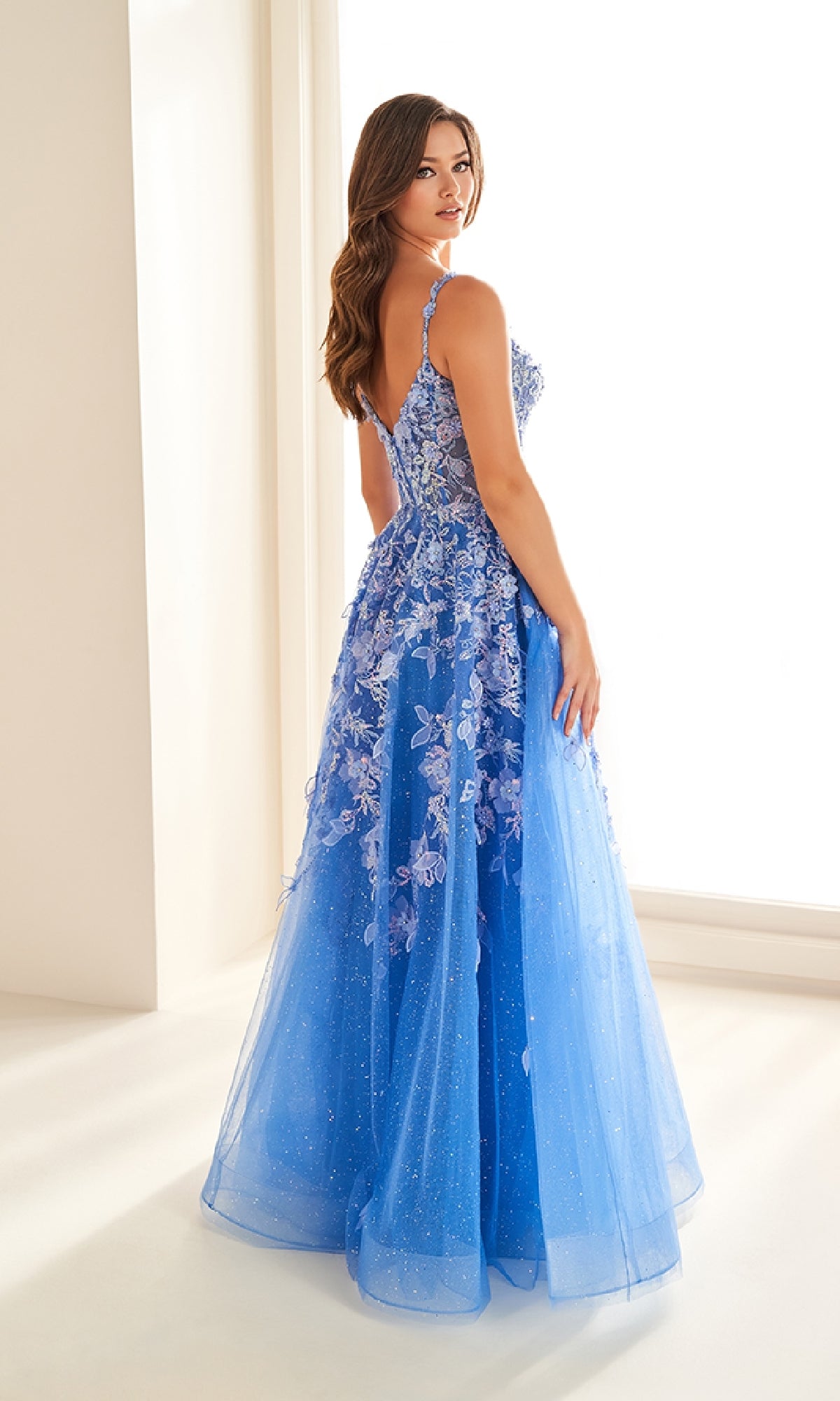 A woman wearing the Ellie Wilde EW36208, a blue, floor-length prom dress with a corset bodice and floral appliqués, stands indoors and looks over her shoulder.