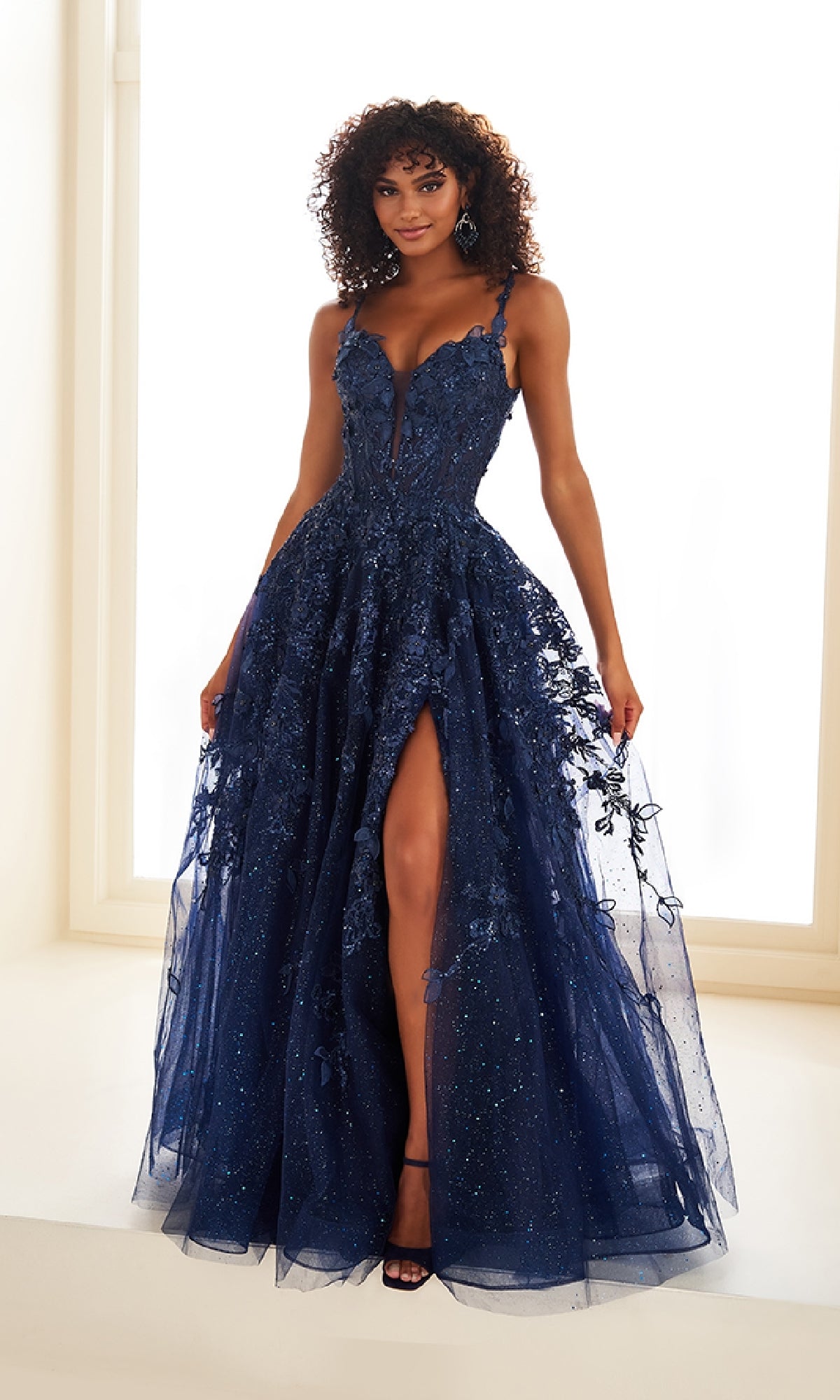 A woman wears the Ellie Wilde EW36208 long prom dress—a navy blue, sleeveless gown with a corset bodice, deep neckline, high slit, and 3D floral embellishments over a light background.