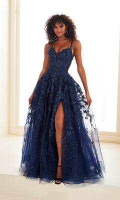 A woman wears the Ellie Wilde EW36208 long prom dress—a navy blue, sleeveless gown with a corset bodice, deep neckline, high slit, and 3D floral embellishments over a light background.