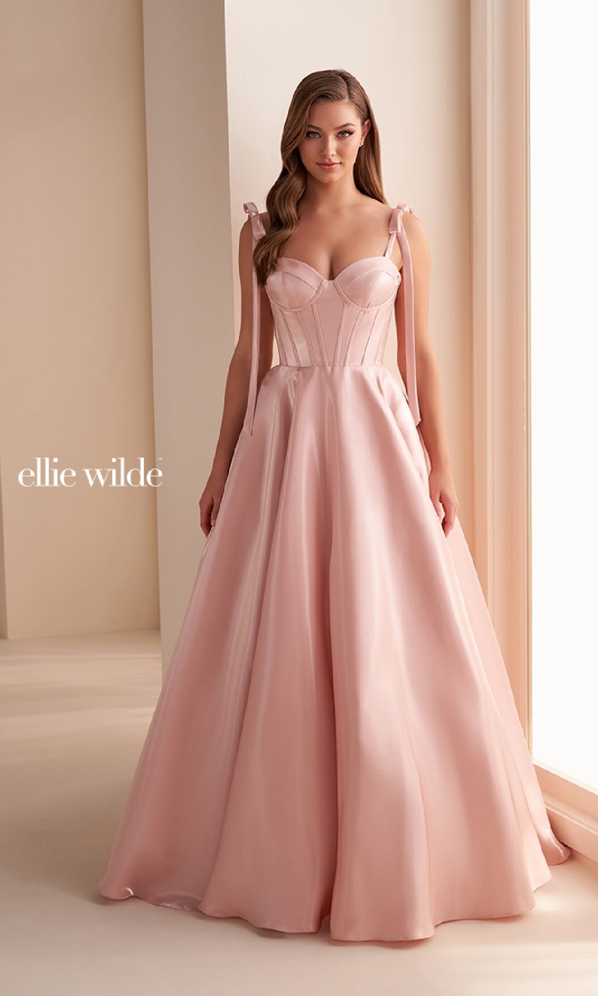 A woman stands indoors in the Ellie Wilde EW36210 Shoulder-Bows Prom Ball Gown—a light pink, floor-length satin A-line dress with a fitted bodice and tie straps. The background is minimal and neutral, with 