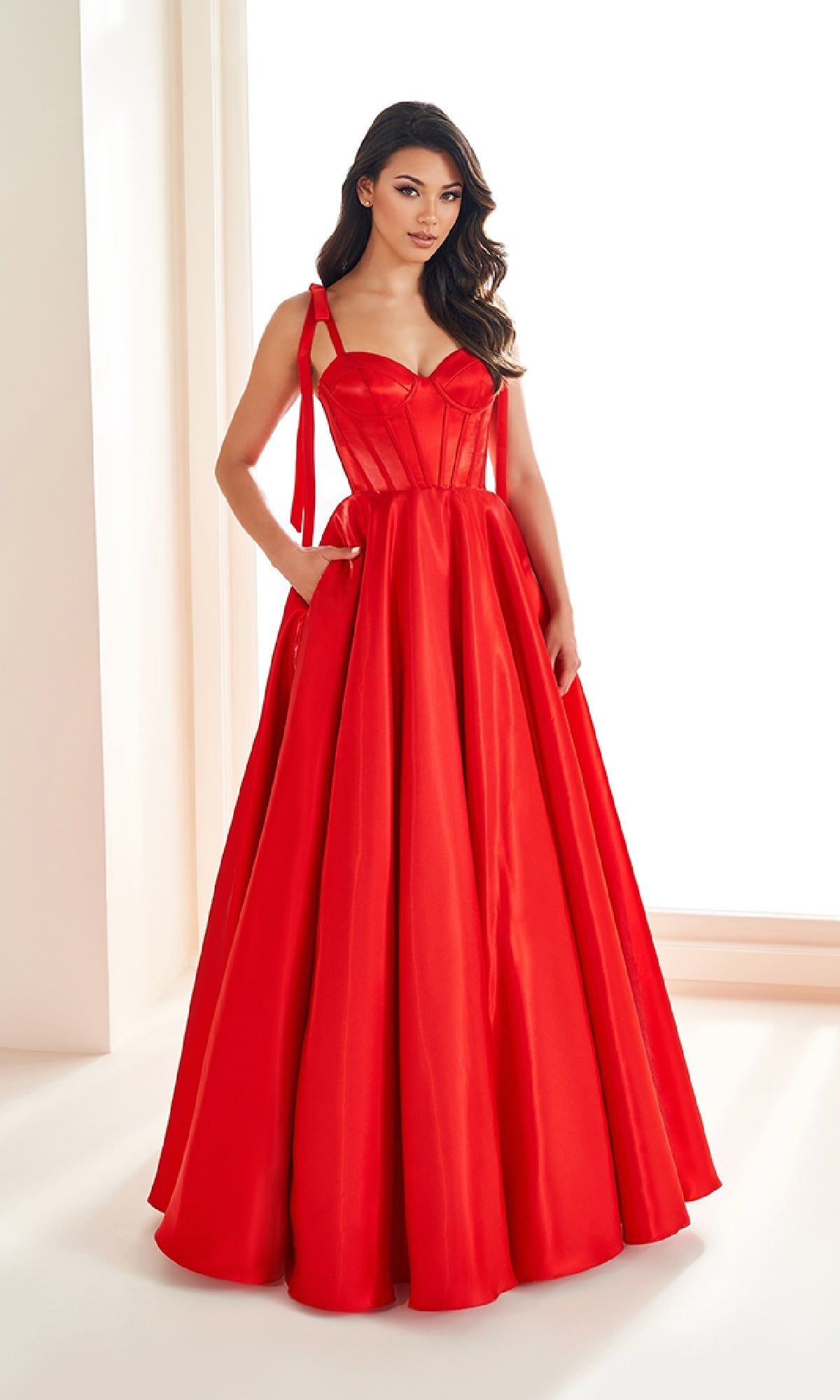 A woman stands indoors wearing the Shoulder-Bows Prom Ball Gown: Ellie Wilde EW36210, a bright red, floor-length prom dress with a fitted bodice and elegant shoulder bows.