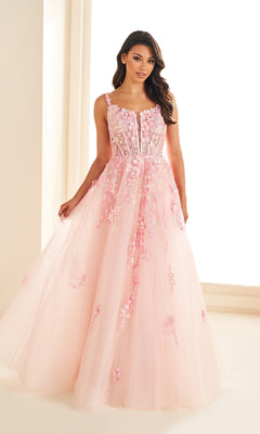 A woman stands in a well-lit room wearing the Ellie Wilde EW36213, a sleeveless, floor-length pink prom dress featuring floral lace appliques and a corset bodice.