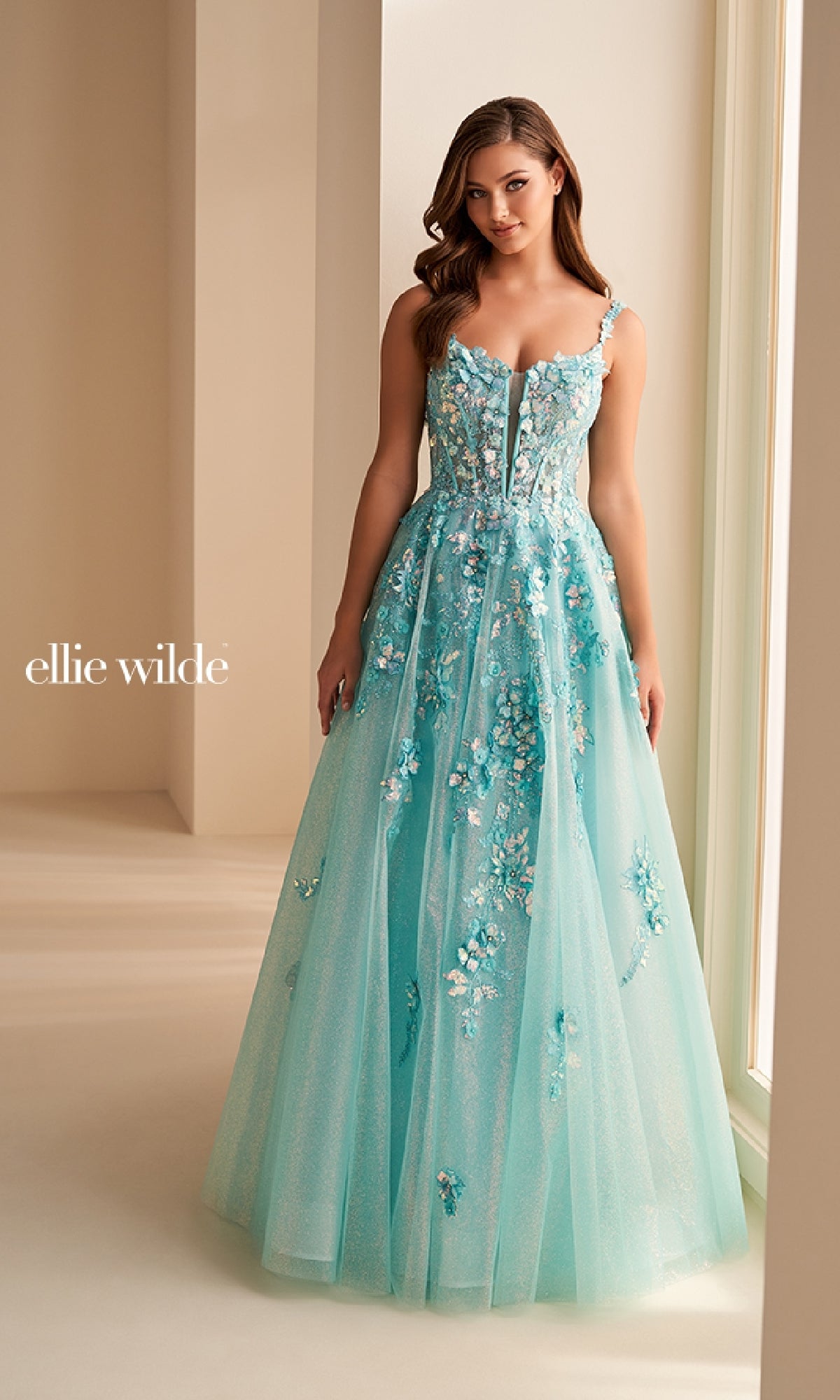 A woman poses indoors in the Ellie Wilde EW36213 long prom dress, showcasing a floor-length turquoise glitter tulle gown with floral appliqués, a sheer corset bodice, and a full tulle skirt. 