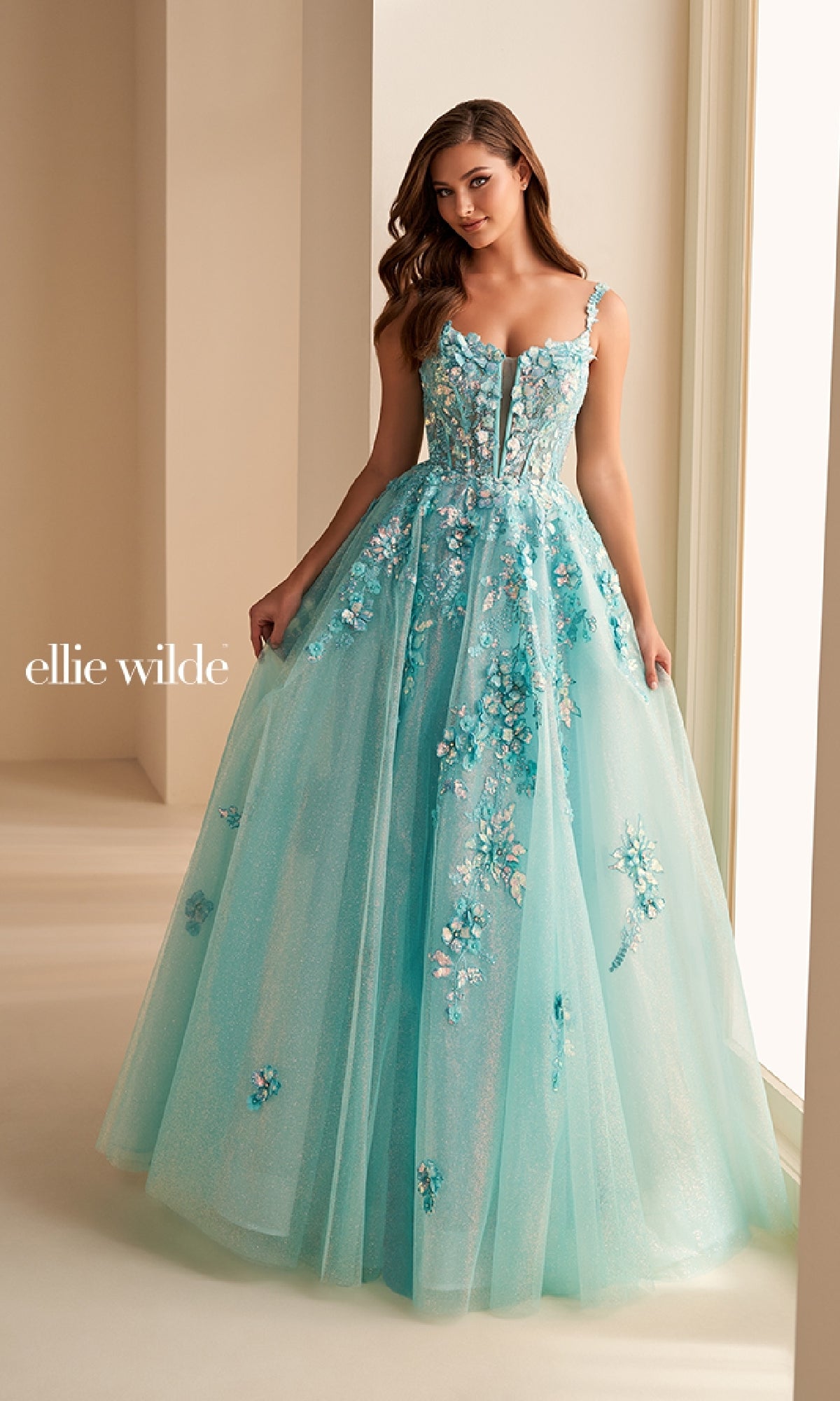 A woman wears the Ellie Wilde EW36213 long prom dress: a light blue, floral-embroidered gown with floral lace appliques, a fitted corset bodice, and a flowing tulle skirt. She is indoors and the 