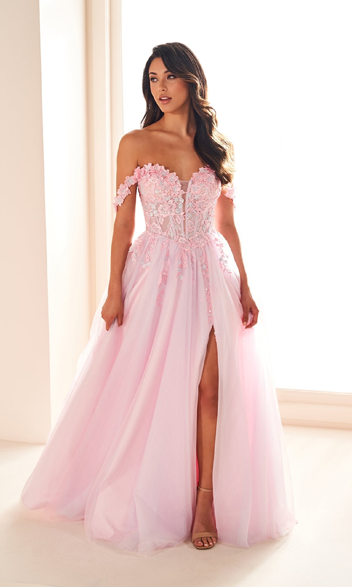Woman wearing the Ellie Wilde EW36214 long prom dress—an off-the-shoulder, light pink a-line tulle gown with floral lace applique corset bodice and high slit—standing by a window indoors.