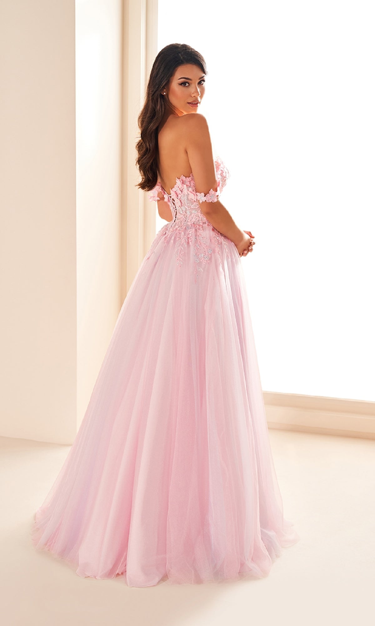 A woman stands in a well-lit room wearing the Ellie Wilde EW36214 long prom dress, featuring an a-line tulle skirt and a lace applique corset bodice with floral detailing.