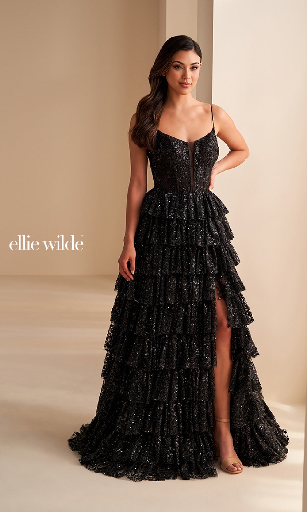 A woman stands in a room wearing the Ellie Wilde EW36216, a sparkly black tiered long prom dress with a corset bodice and front slit. The text 