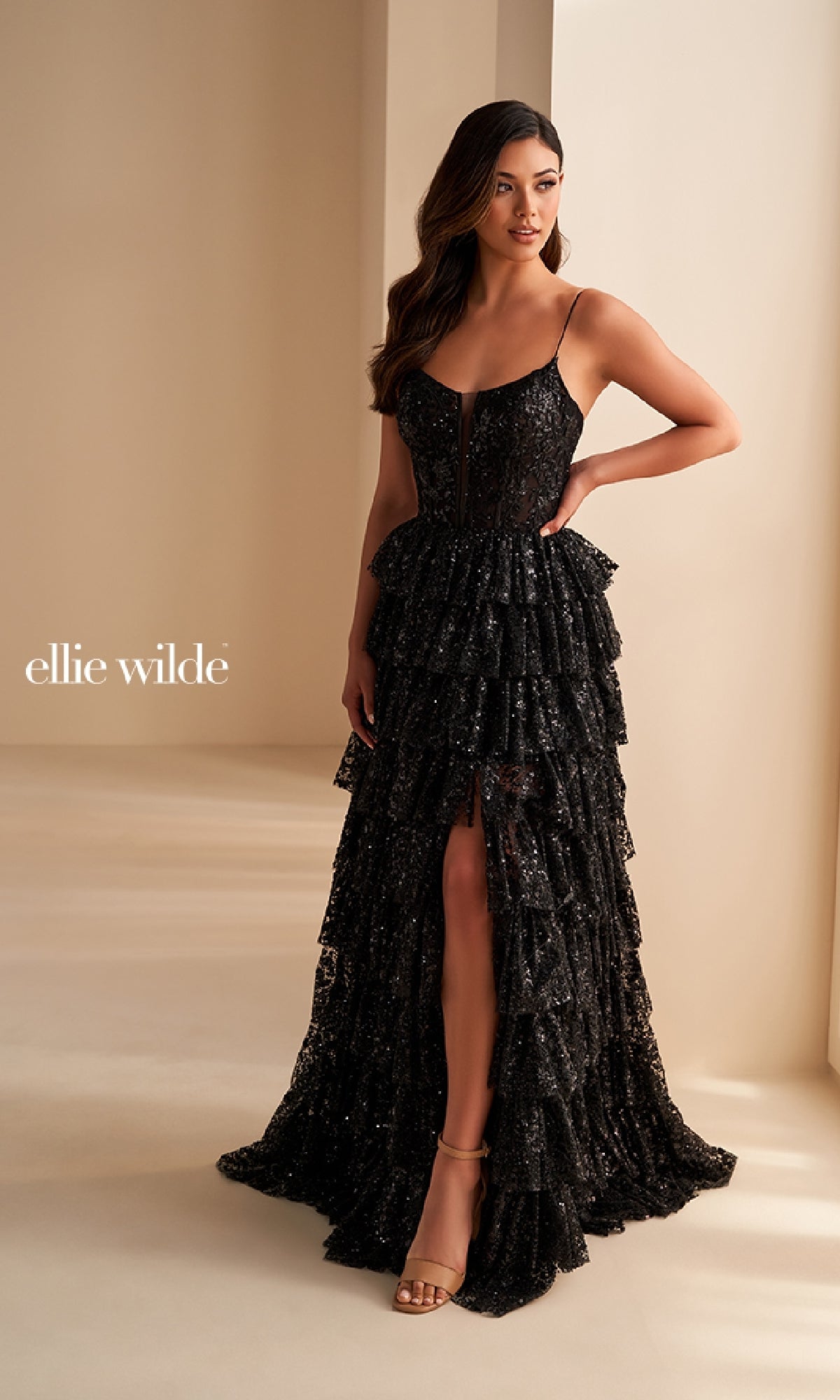 A woman stands in a neutral-toned room wearing the Ellie Wilde EW36216 long prom dress—black with spaghetti straps, a corset bodice, tiered ruffles, and a thigh-high slit. 