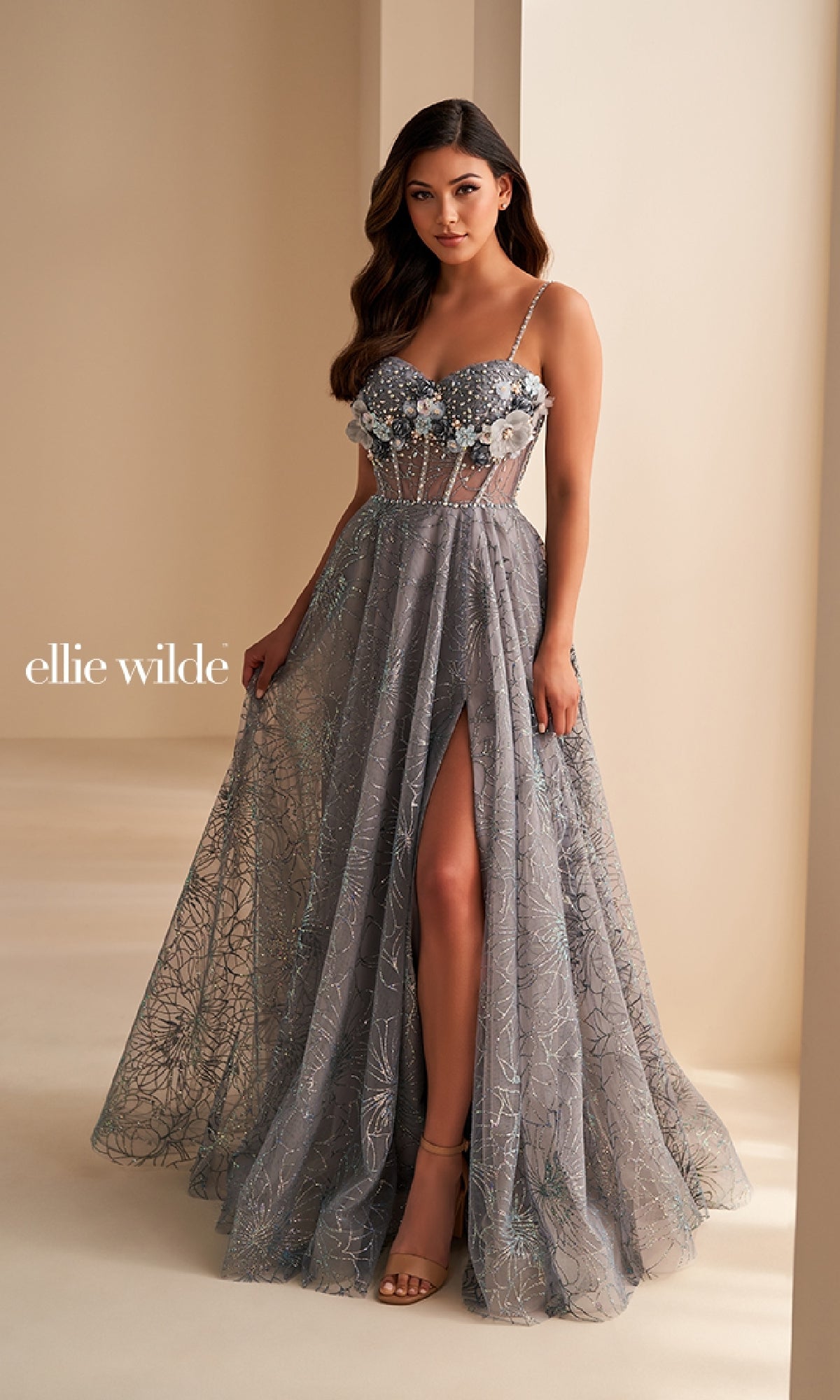 A woman wears the Ellie Wilde EW36218 long prom dress, featuring a sheer silver-gray embellished gown with 3D floral appliqué, spaghetti straps, a high slit, and a sheer corset bodice, standing indoors against a neutral background.