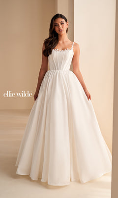 A woman stands indoors wearing the Ellie Wilde EW36221 long prom dress, featuring a white A-line skirt, a fitted sleeveless bodice with delicate straps, subtle pleating, and a simple, elegant design.