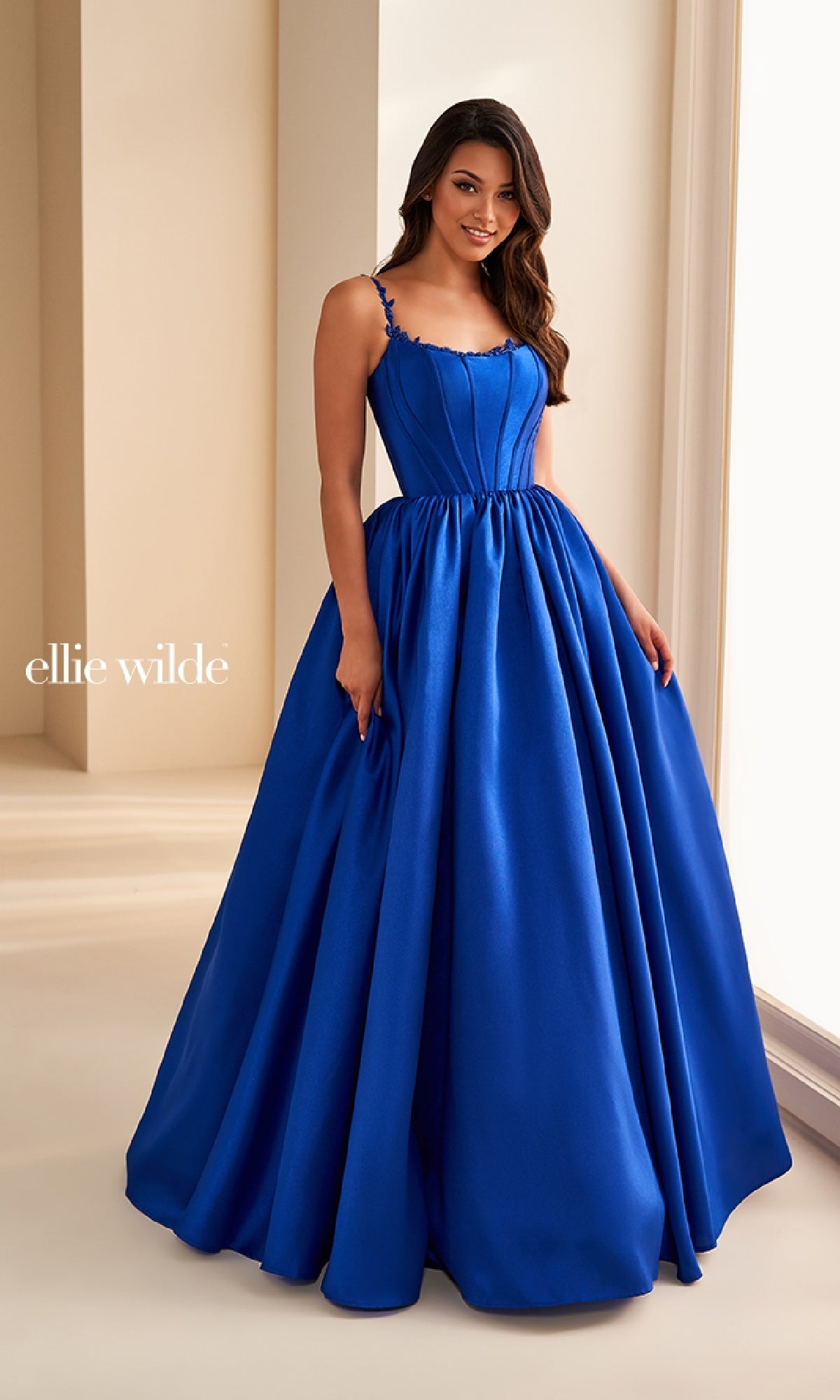 A woman wears the Ellie Wilde EW36221 long prom dress indoors. The royal blue gown has thin straps, a fitted corset bodice, and a full A-line skirt. 
