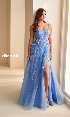 A woman stands by a window in a softly lit room wearing the Ellie Wilde EW36222, a light blue, floor-length prom dress with thin straps, lace applique, thigh-high slit, and fitted corset bodice.