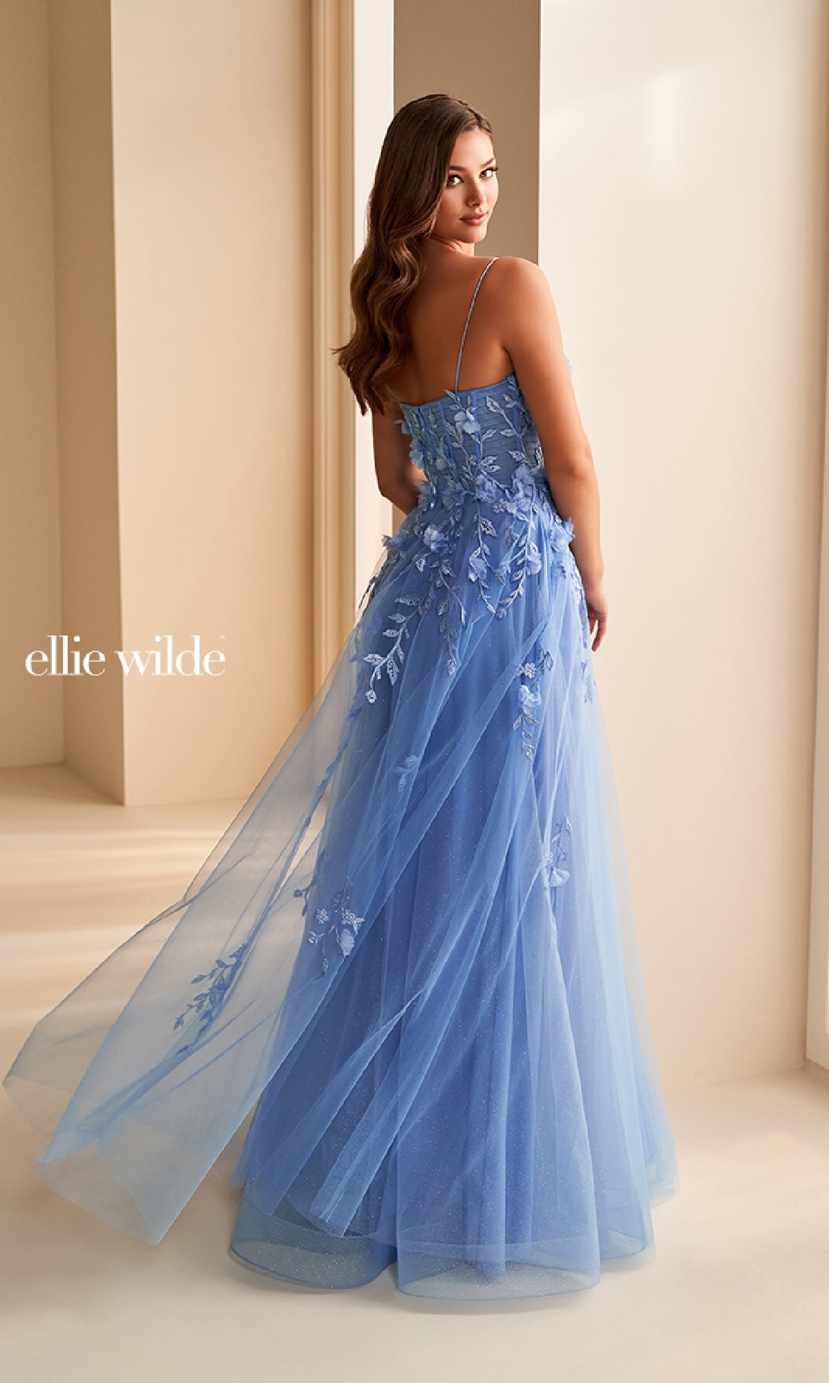 A woman wears the Ellie Wilde EW36222 long prom dress, featuring blue floral embroidery, a floor-length sheer overlay, spaghetti straps, and a fitted corset bodice as she faces away.