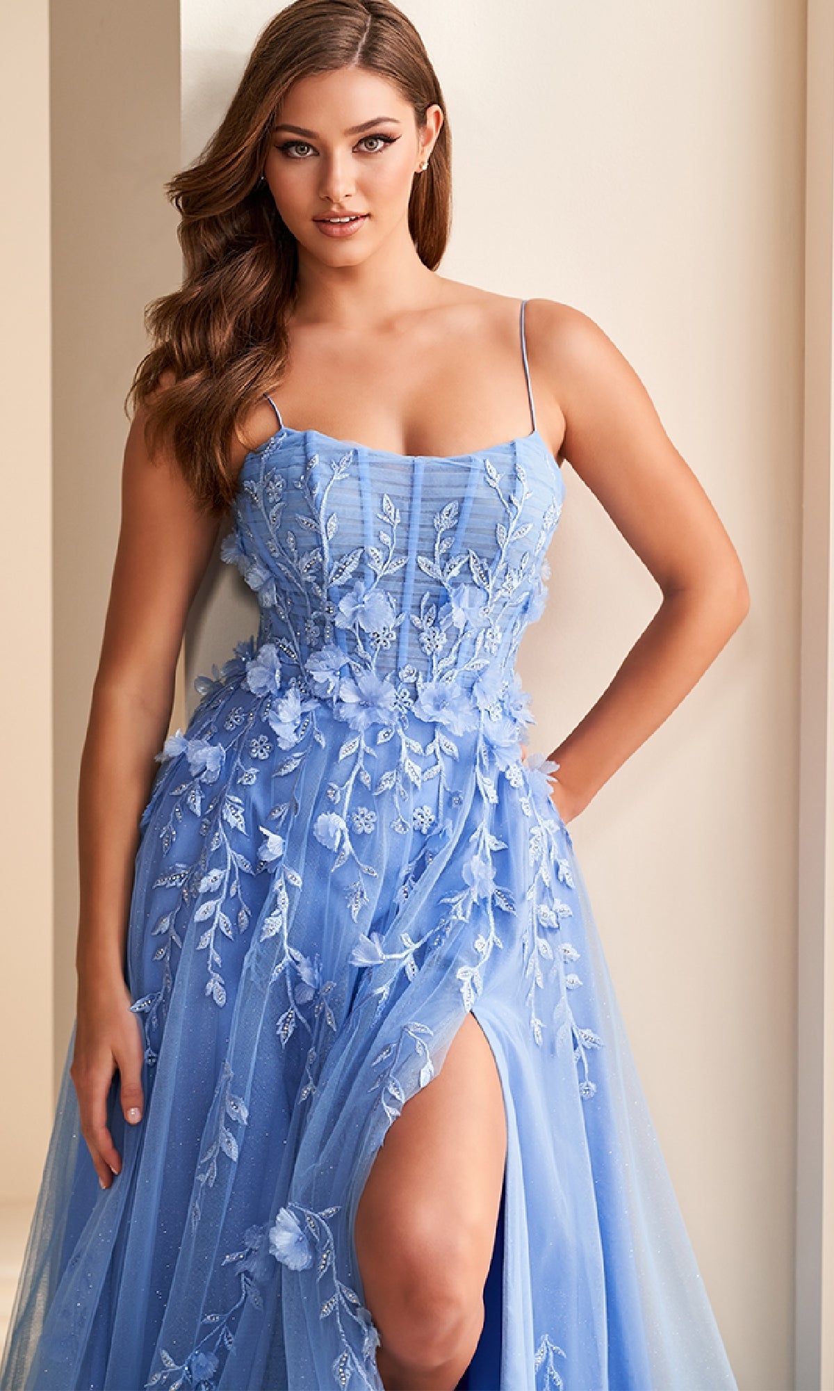 A woman wears the Ellie Wilde EW36222 long prom dress, featuring light blue fabric, delicate floral embroidery, a flattering corset bodice with spaghetti straps, and a thigh-high slit, standing against a neutral background.